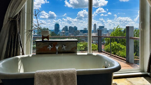 Ein modernes Badezimmer mit einer großen Badewanne, Blick auf einen Stadtpanoramablick durch ein großes Fenster und einem kleinen Tischchen mit einer Vase.