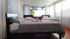 A large bed with a grey quilt, white pillows, and a red accent pillow. There is a window with white curtains and a view of trees outside.