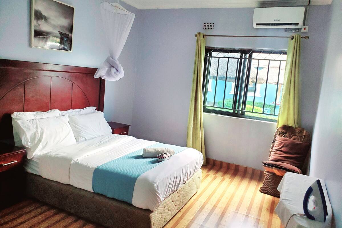 A hotel room with a bed, a window with curtains, an air conditioning unit, and a painting on the wall.