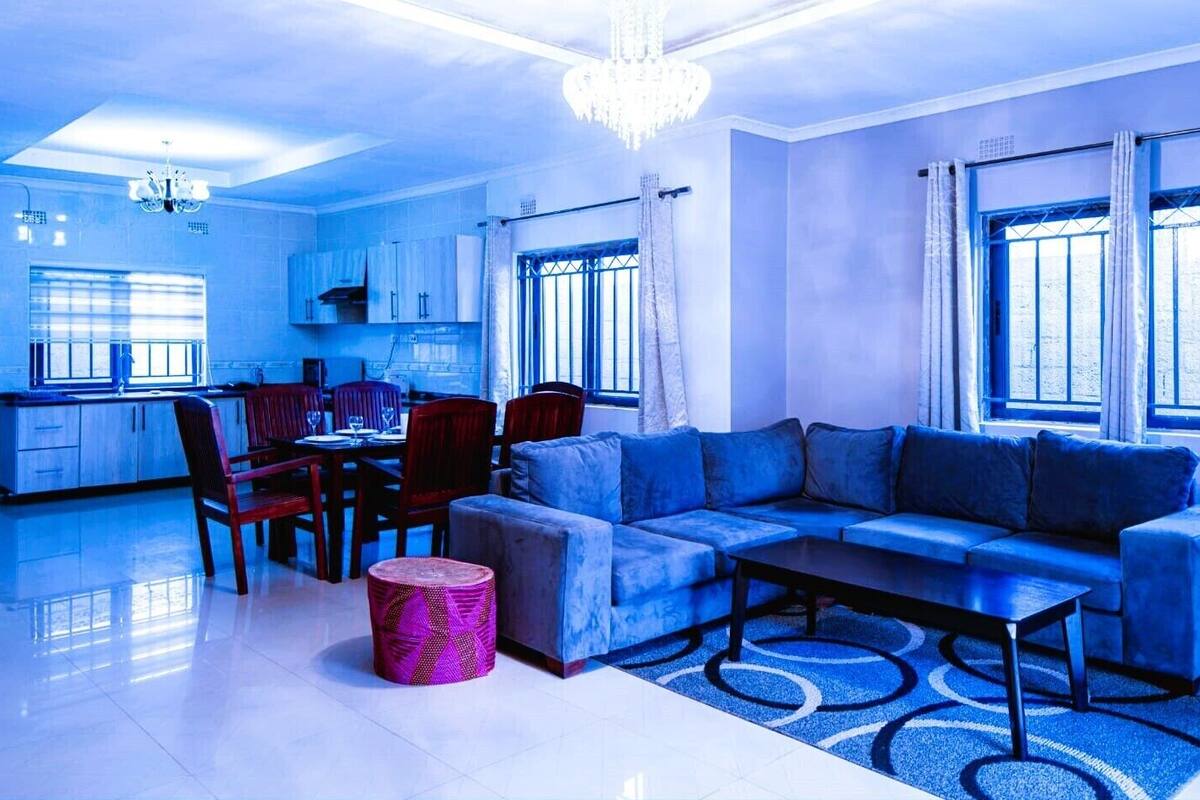 A modern living room with a blue sofa, a dining table with chairs, a kitchen area, and large windows with curtains.