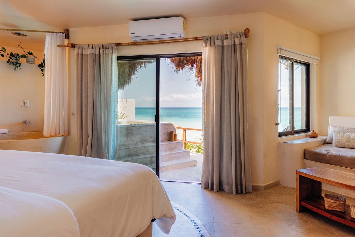 A hotel room with a bed, a seating area with a view of the beach, and a balcony with a railing.