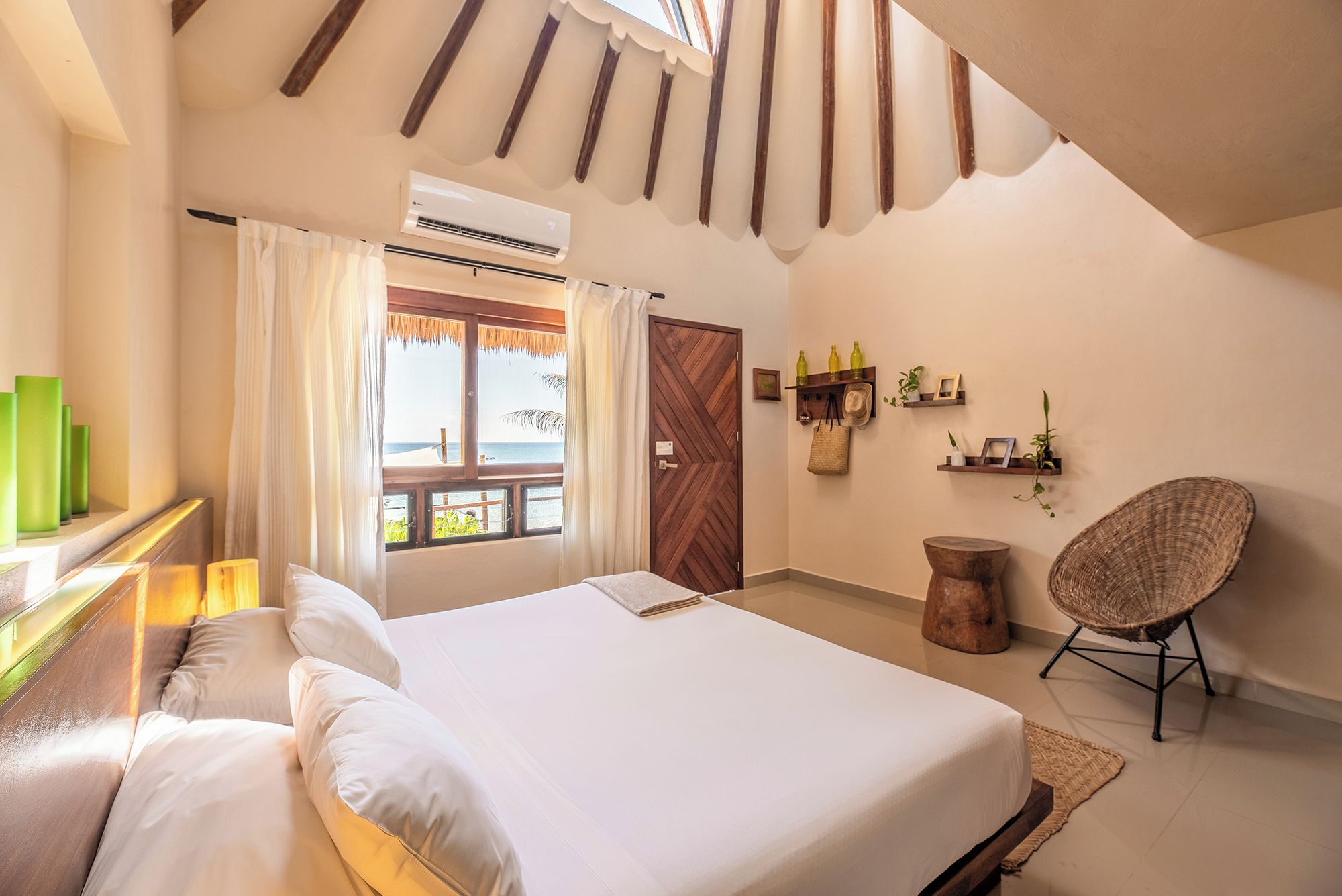 A bedroom with a bed, a chair, a shelf with bottles and a hat, and a view of the sea through a window.