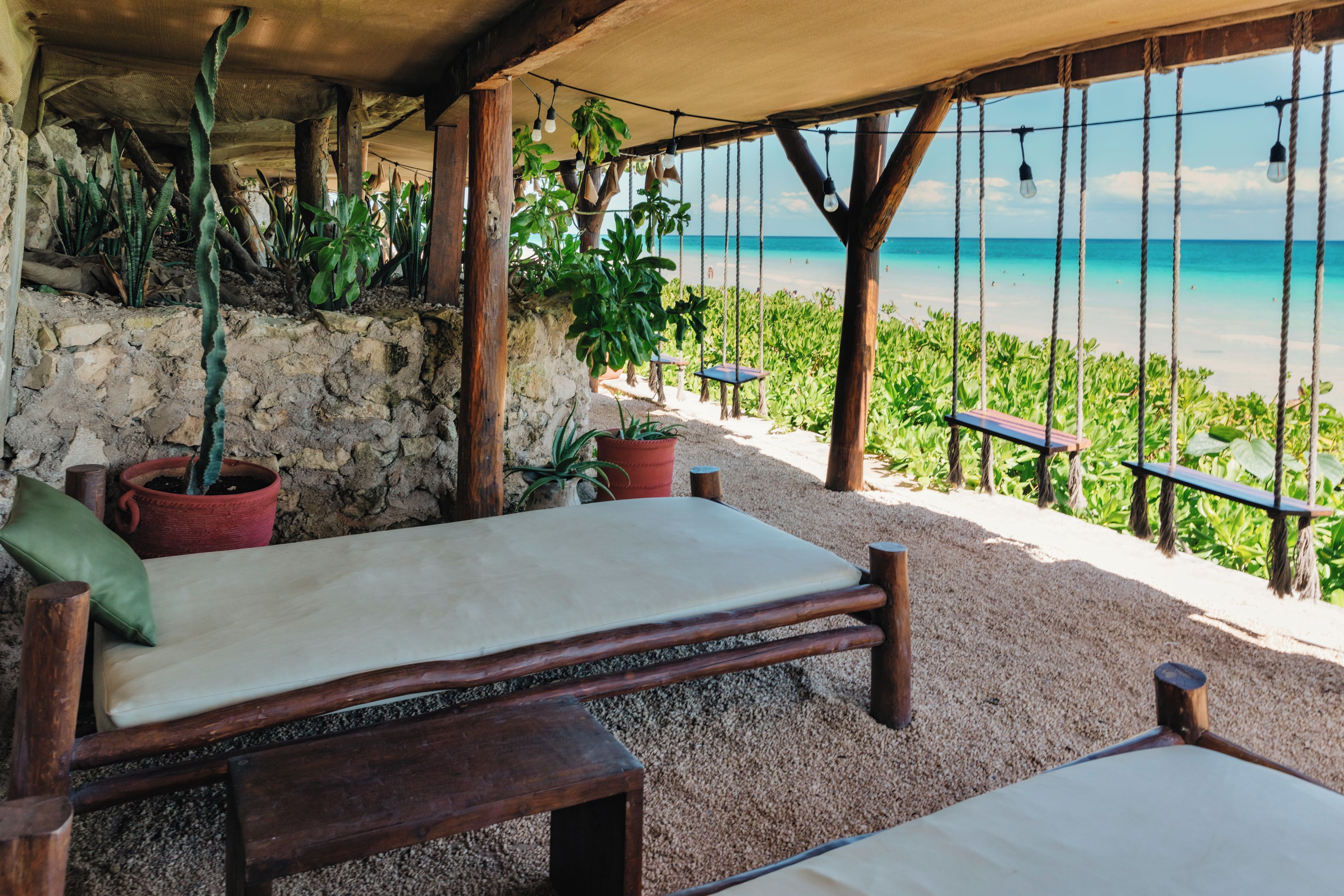 A wooden bench with a cushion, surrounded by potted plants, overlooking a beach with a clear view of the ocean.