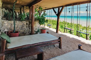 A wooden bench with a cushion, surrounded by potted plants, overlooking a beach with a clear view of the ocean. - Mezzanine by La Zebra, an SLH Hotel (Tulum)