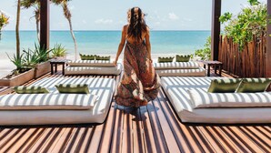 Une femme se promène sur une terrasse en bois, où se trouvent des chaises longues et des coussins, avec vue sur une plage et l’océan.