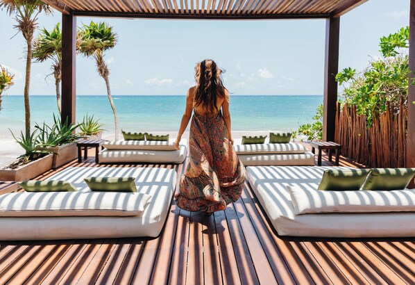 Una mujer camina sobre una terraza de madera con sillas reclinables y cojines, con vistas a una playa y al océano.