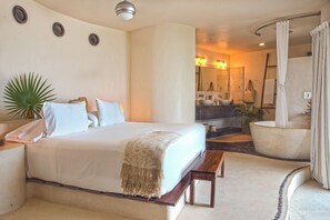 A bedroom with a large bed, bedside tables, a bench, a potted plant, and a bathroom with a bathtub, sink, and mirror. - Mezzanine by La Zebra, an SLH Hotel (Tulum)