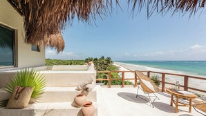 Un balcon donnant sur une plage, un toit de chaume et une chaise en osier.
