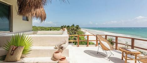 A balcony with a view of a beach, a thatched roof, and a wicker chair.