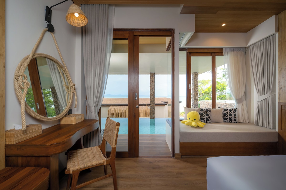 Een kamer met een houten bureau, een stoel, een grote spiegel, een bed met uitzicht op zee, en een balkon met een zwembad.