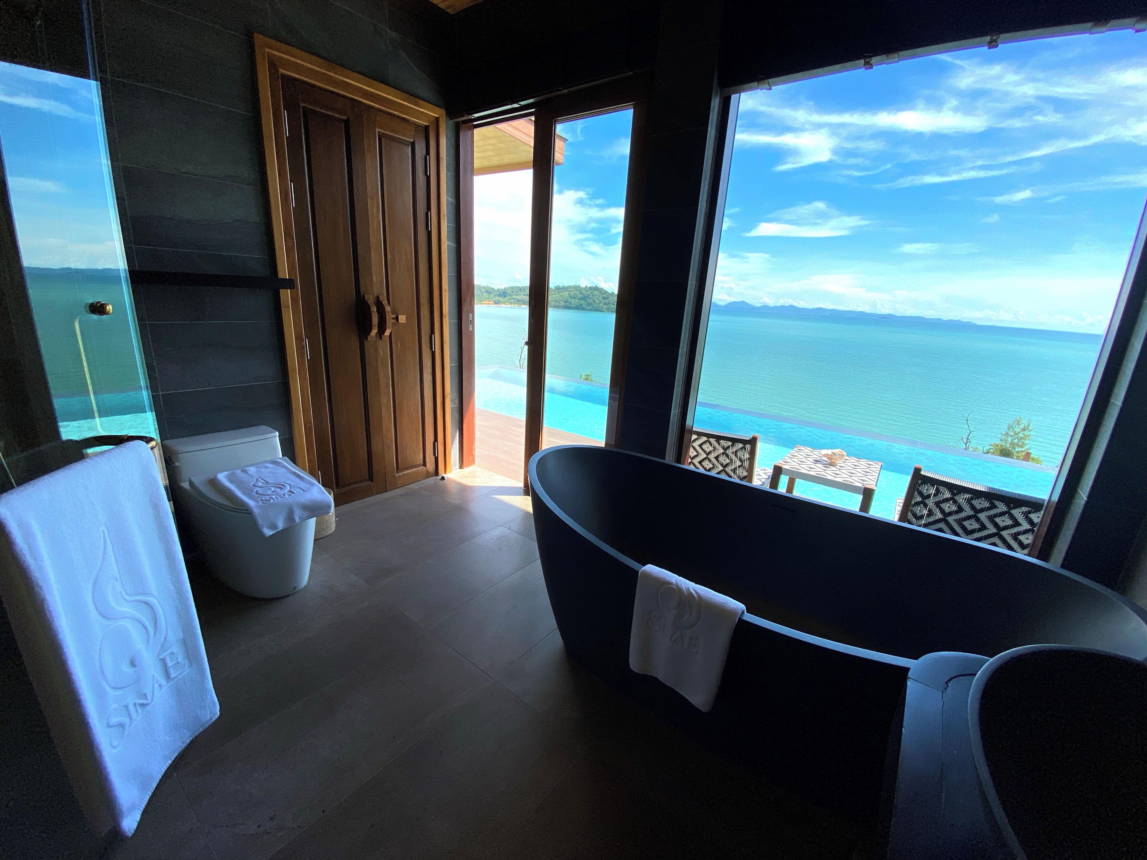 Een badkamer met een bad, toilet en douche, met een groot raam en uitzicht op zee.