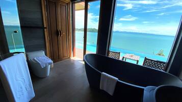 Een badkamer met een bad, toilet en douche, met een groot raam en uitzicht op zee.