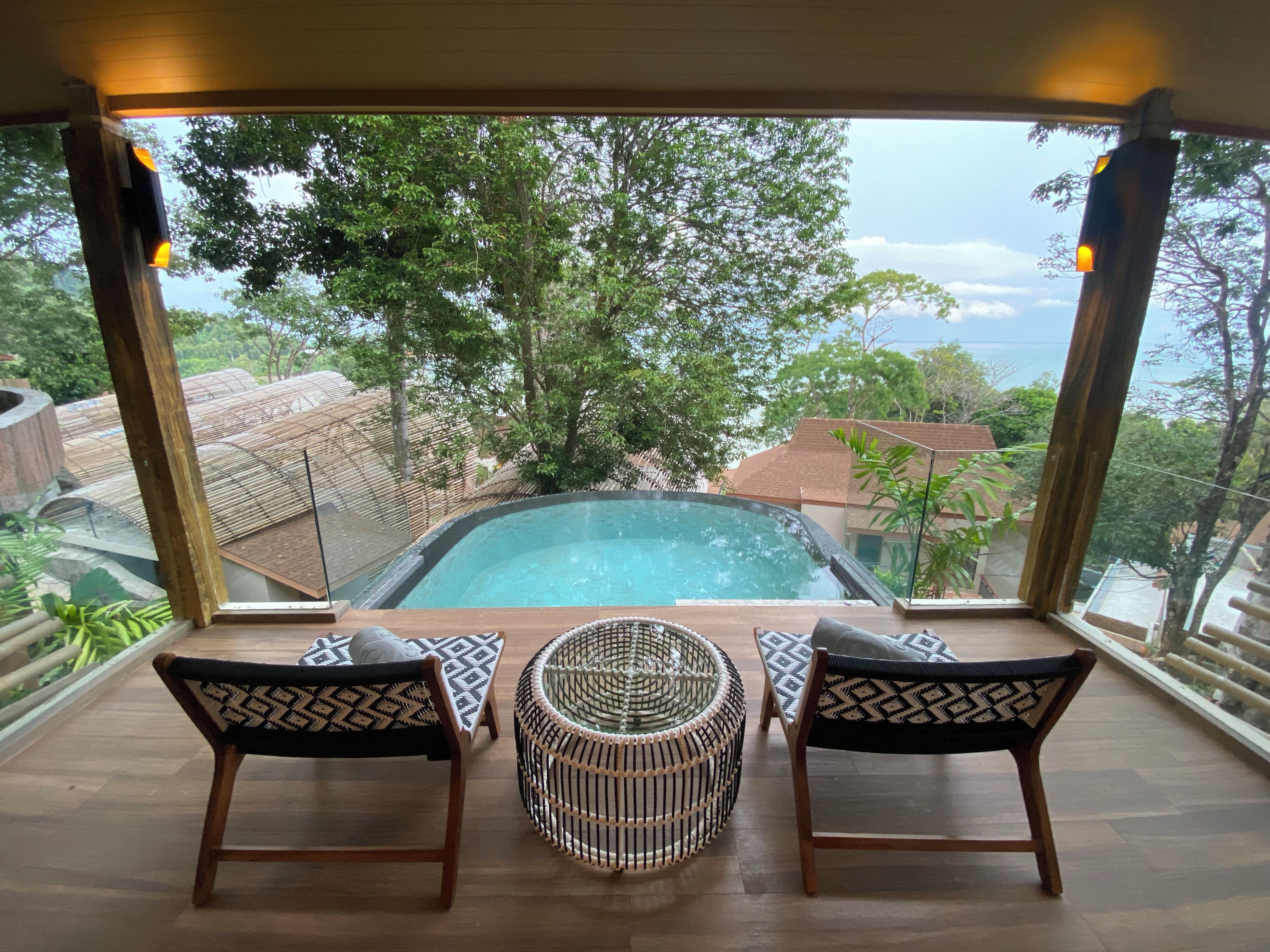 A pool area with two chairs and a table, surrounded by glass walls offering a view of trees and a distant landscape.