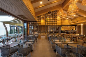 Interior de restaurante con techos altos, vigas de madera y diferentes opciones de asientos, incluyendo mesas y sillas de mimbre.