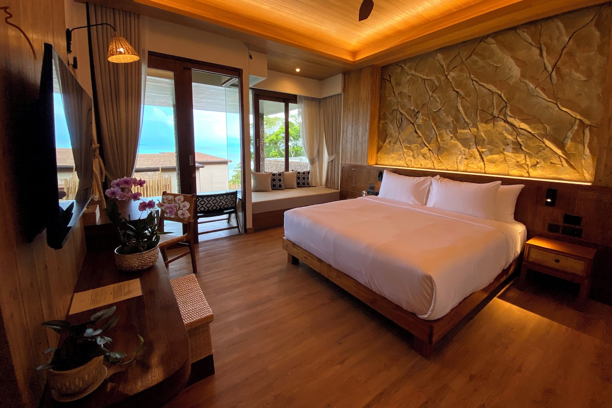A hotel room with a large bed, a ceiling fan, a view of the ocean, and a stone wall.
