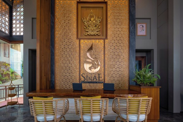 Una zona d'entrada amb un taulell de recepció de fusta, dues cadires, una planta en test i una paret decorativa amb un emblema daurat i el nom 'SINAE PHUKET LUXURY HOTEL'.