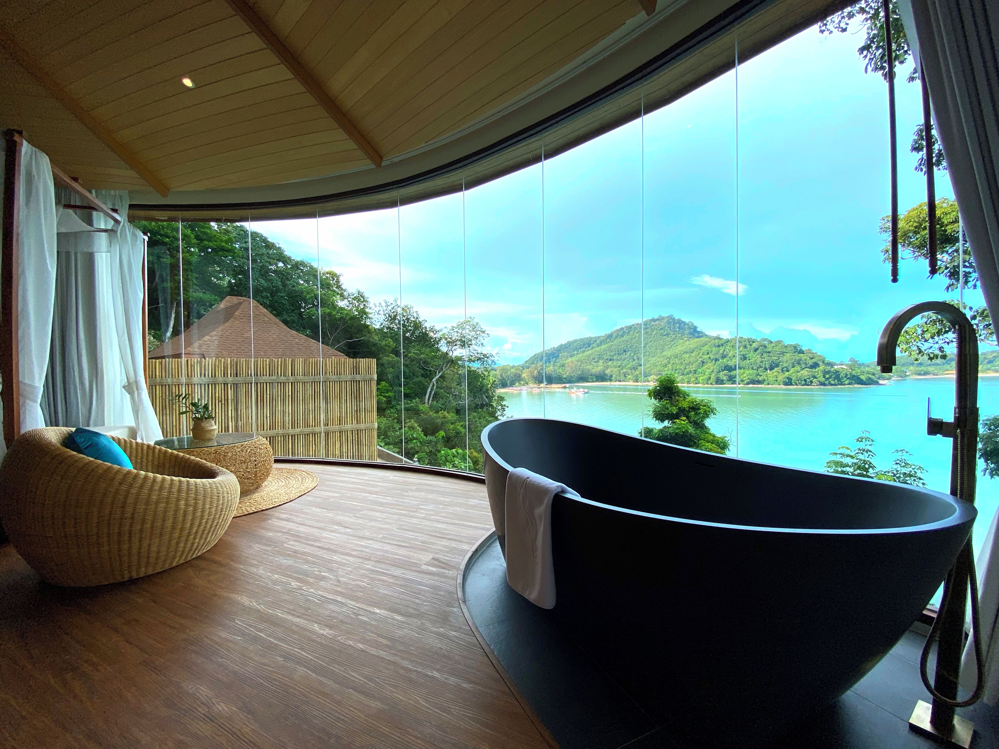 A modern bathroom with a freestanding bathtub, wicker furniture, and a large window offering a scenic view of a lake and mountains.