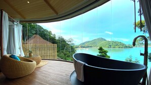 Une salle de bain moderne avec une baignoire sur pieds, des meubles en osier et une grande fenêtre offrant une vue panoramique sur un lac et des montagnes.