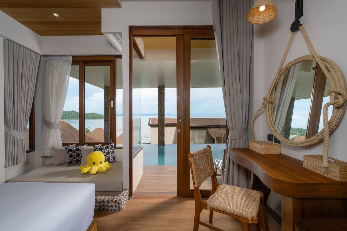 Een slaapkamer met een bed, een stoel, een houten bureau, een grote spiegel en uitzicht op zee.