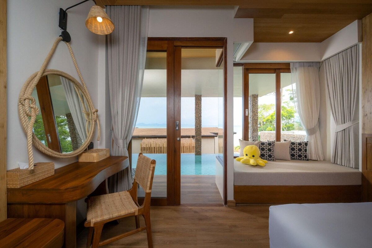 Une pièce avec un bureau en bois, une chaise, un lit avec un doudou jaune, un grand miroir et une vue sur une piscine et une plage.