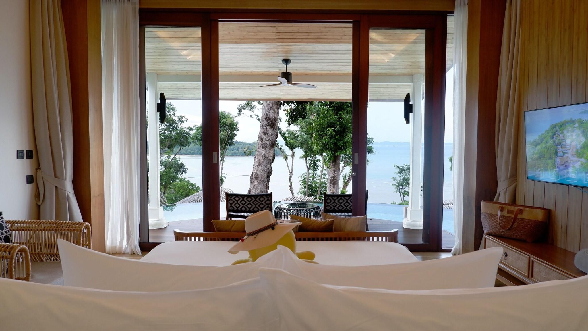 Une chambre d’hôtel avec un lit, une télévision, un fauteuil en osier, une table en bois et une vue sur la mer.