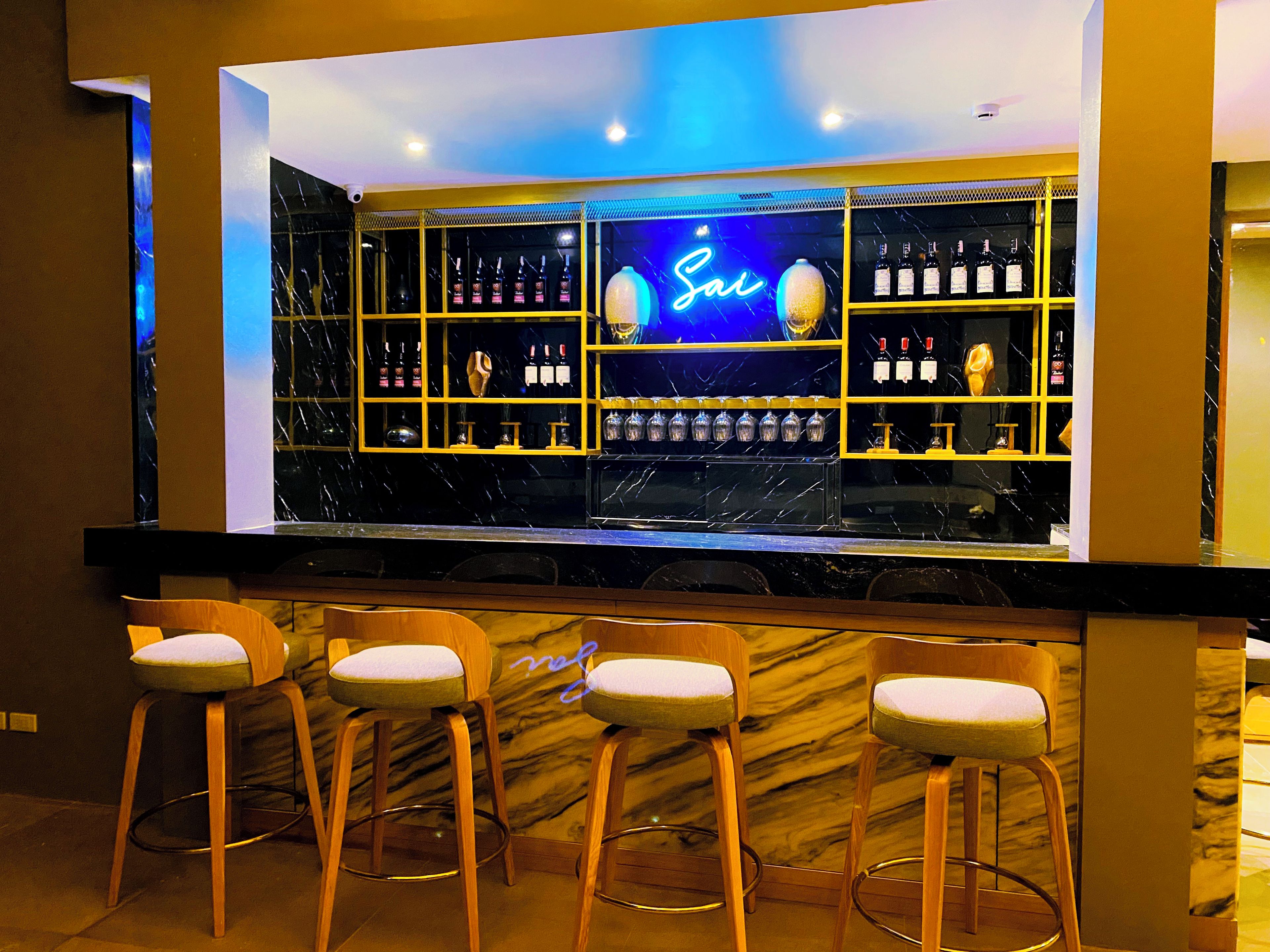 Zona de bar con un letrero de neón, estantes llenos de botellas y tres taburetes.