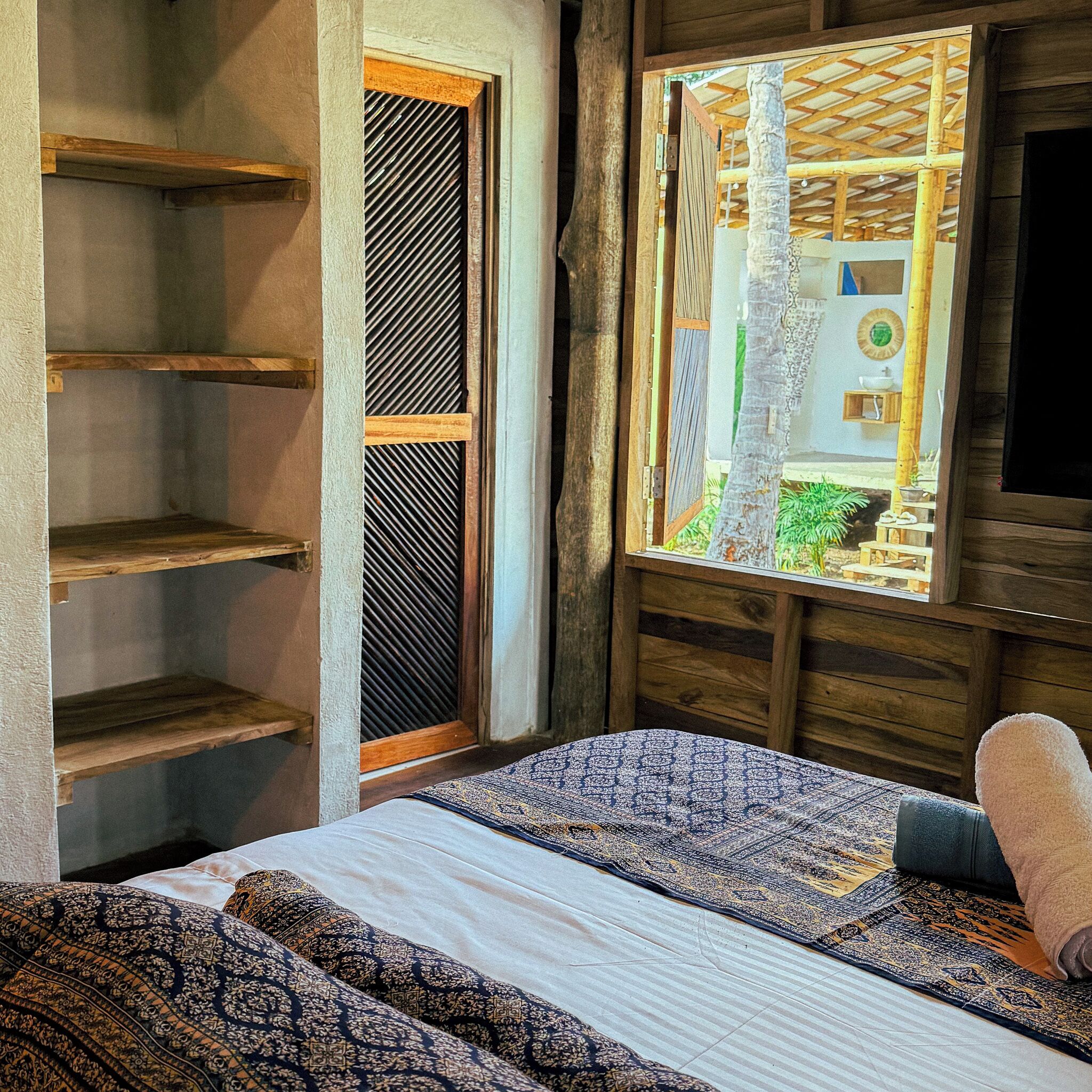 A room with a bed, a wooden shelf unit, and a window with a view of an outdoor area.