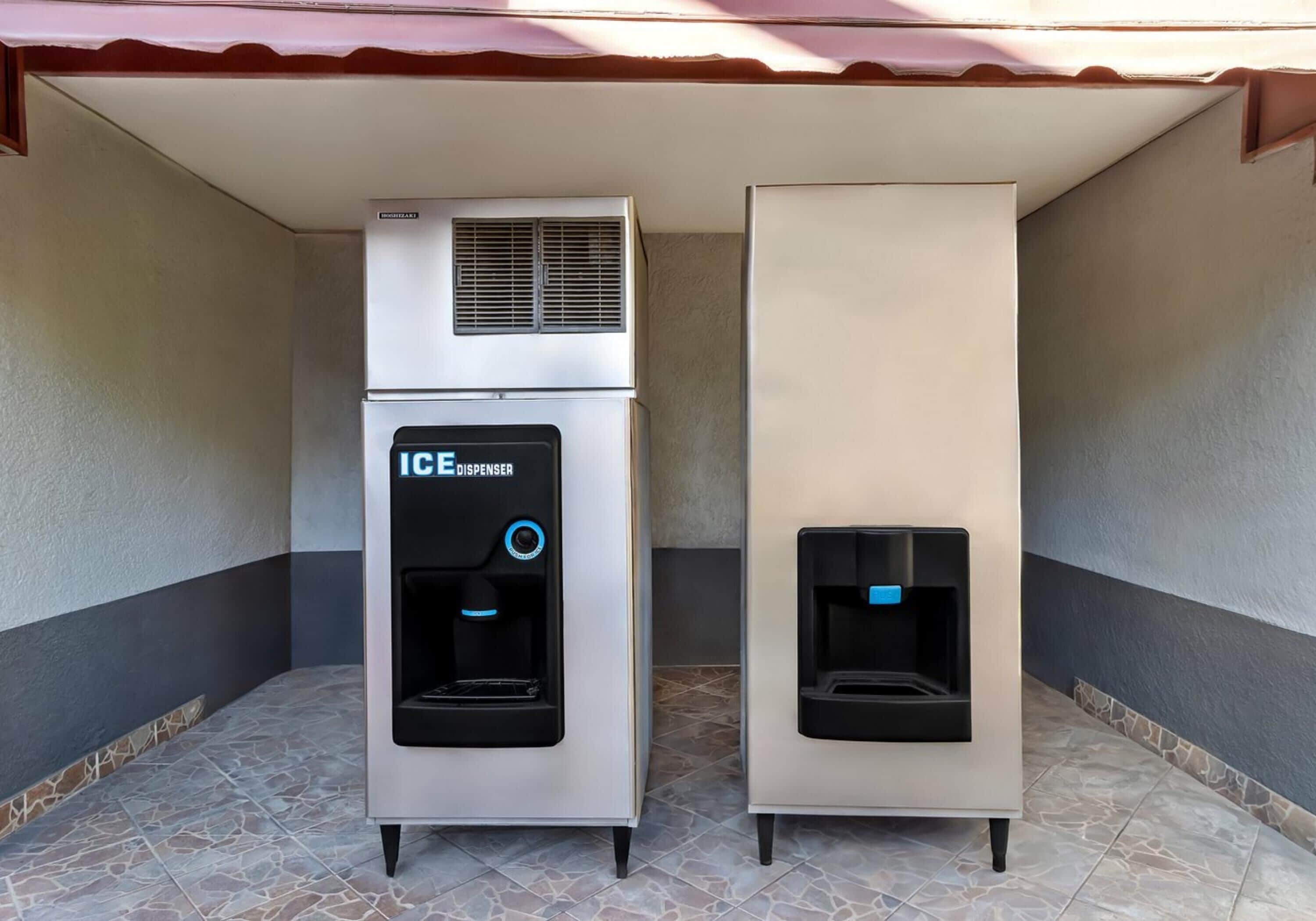 Two ice dispensers, one with a freezer and one without, on a tiled floor under a roofed area.