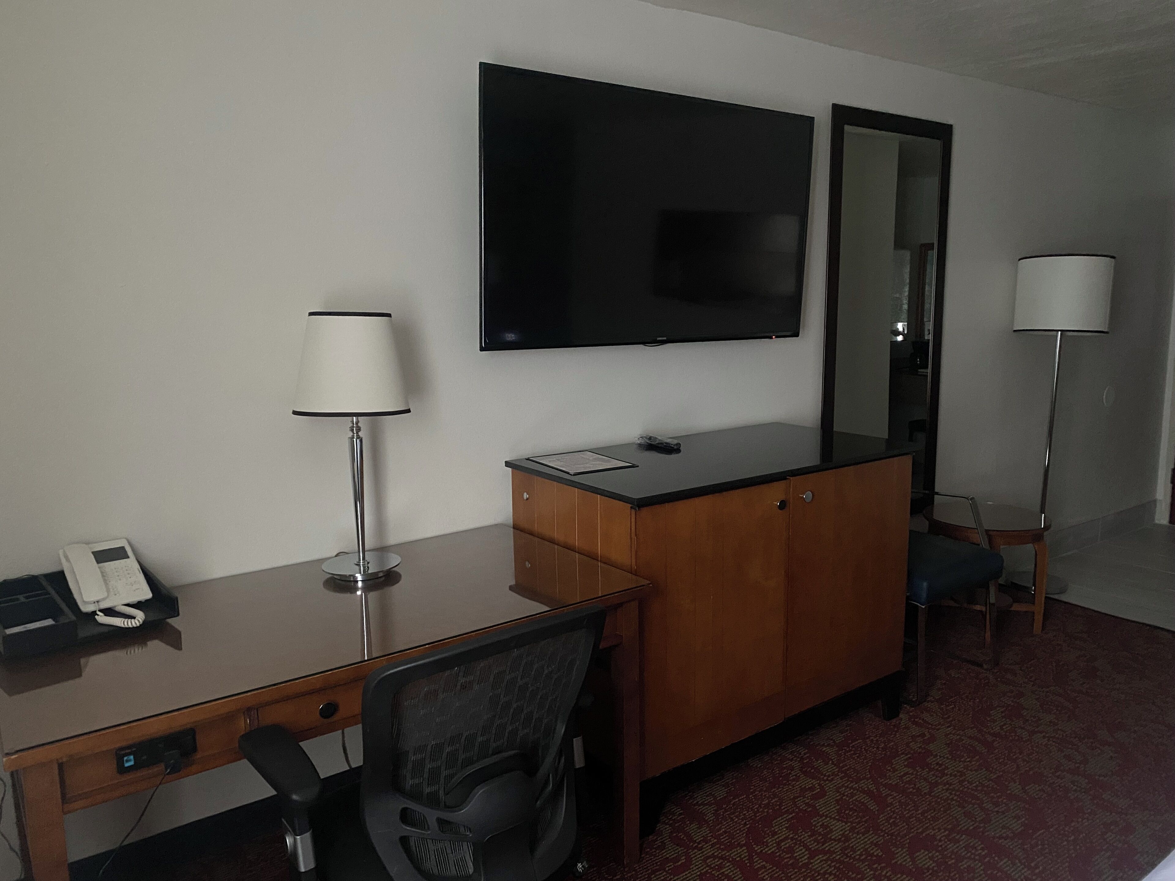 A hotel room with a desk, chair, lamp, telephone, and a flat-screen TV mounted on the wall.