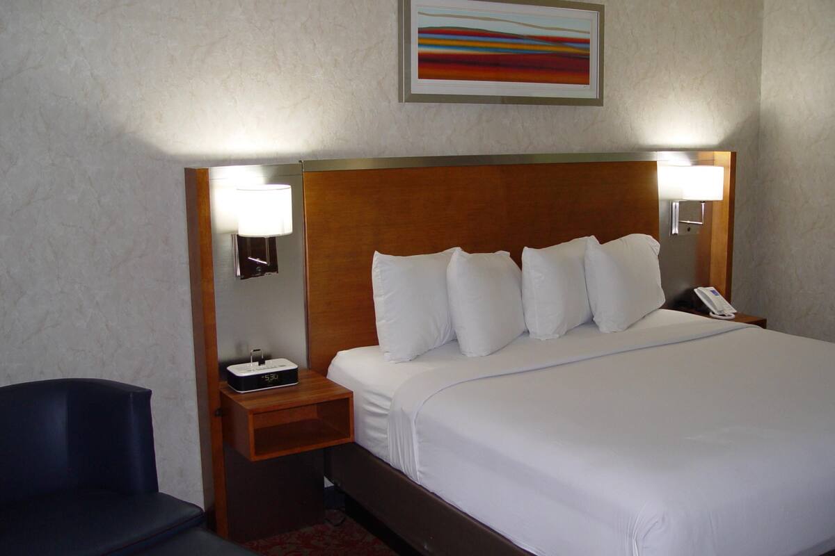 A hotel room with a bed, bedside lamps, a phone, a chair, and a painting on the wall.