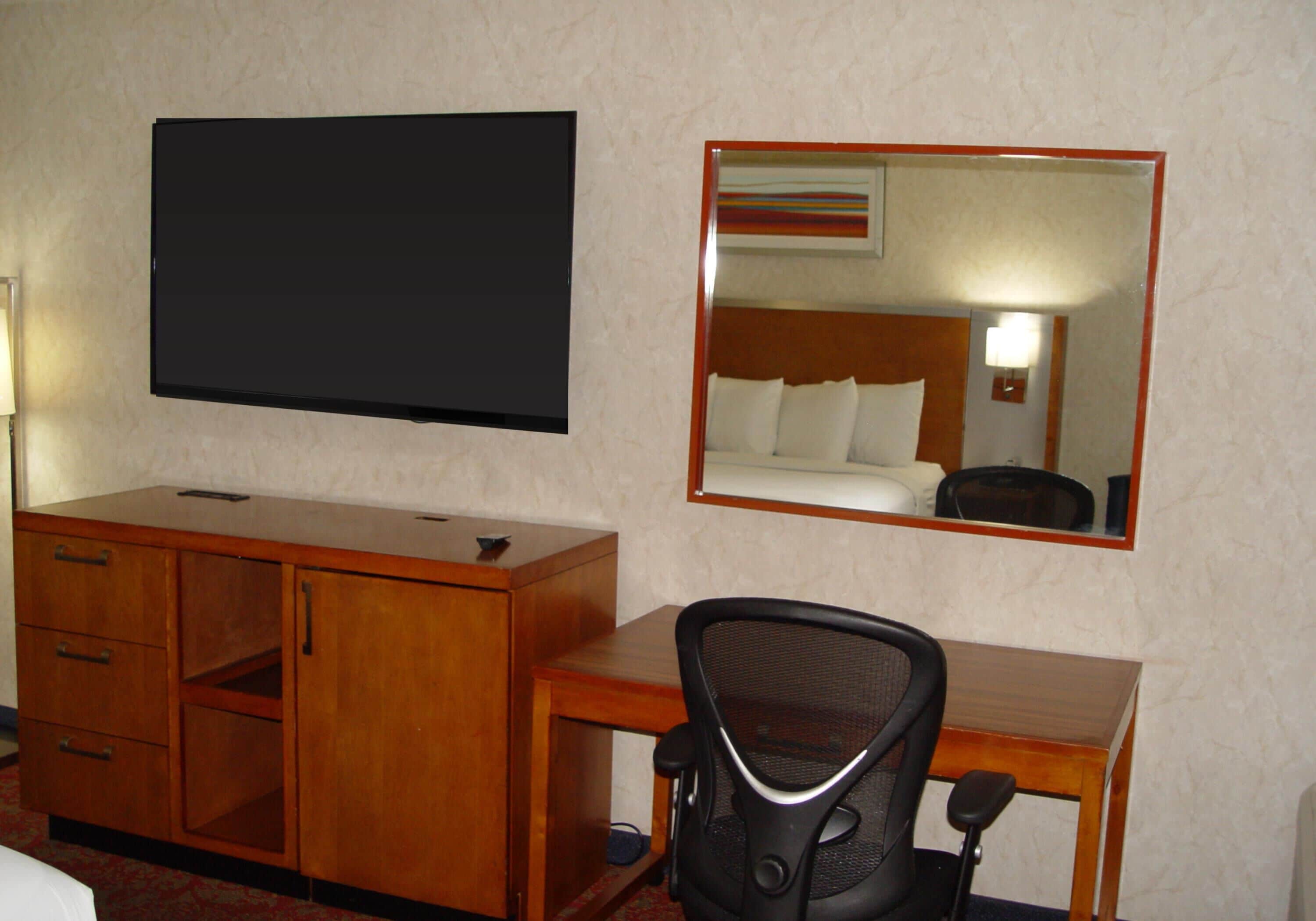A hotel room with a flat-screen TV, a wooden desk, a chair, a mirror, and a bed.