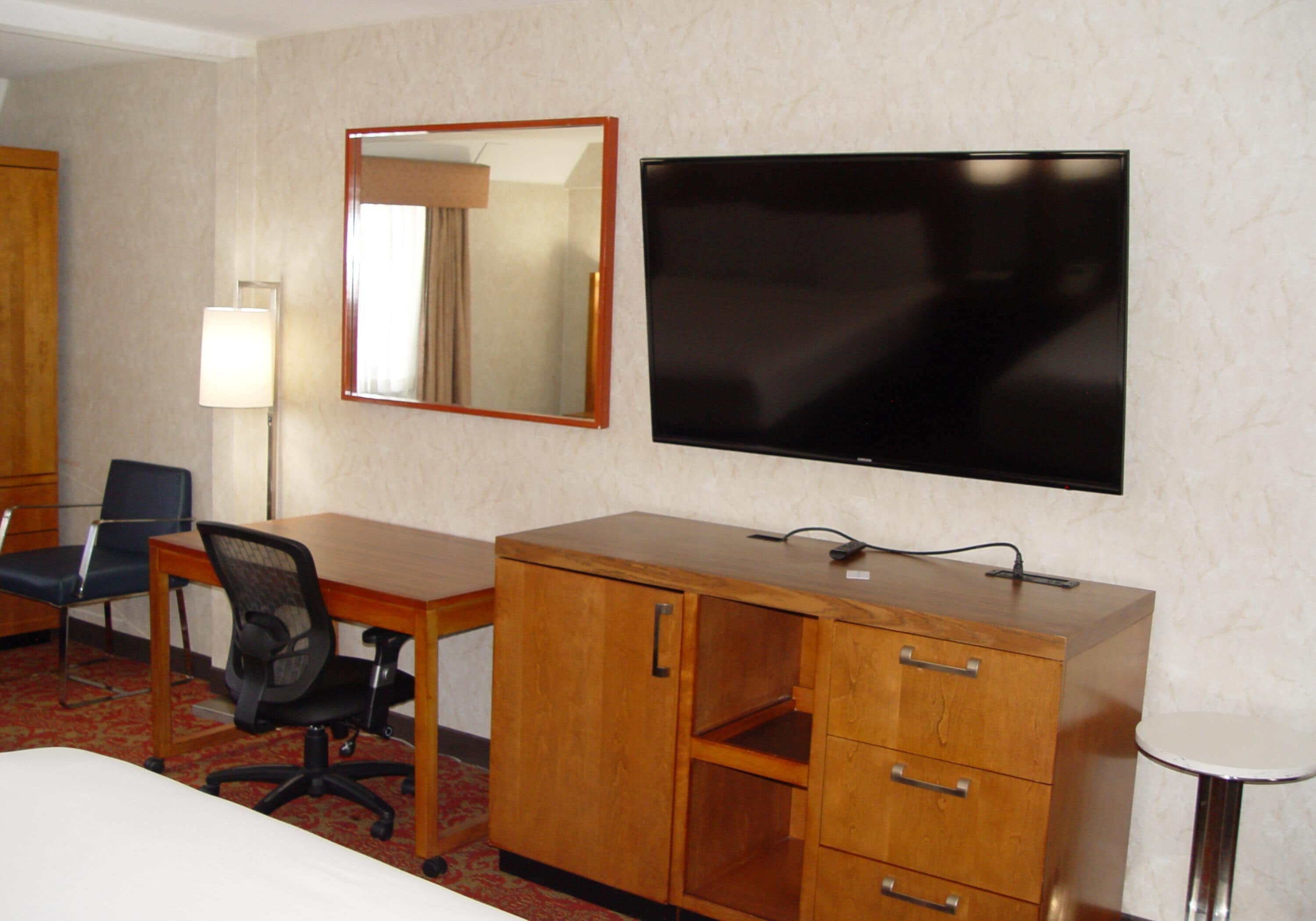 Una camera d'albergo con una scrivania in legno, una sedia, una televisione, uno specchio e un tavolino rotondo.