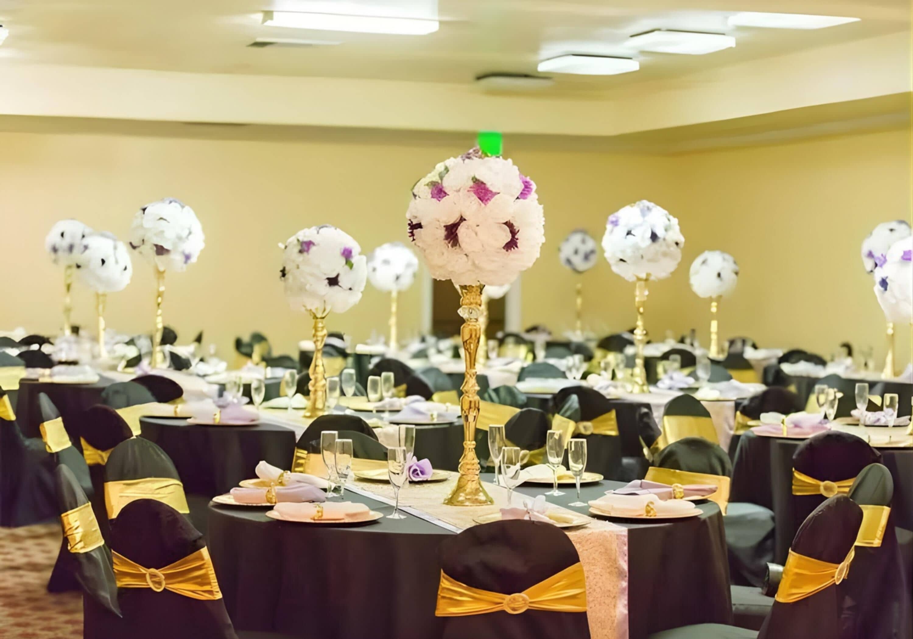 A banquet hall with round tables, black chairs with gold sashes, and floral centerpieces on tall stands.