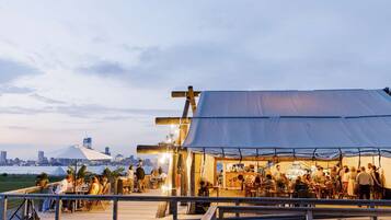 Un restaurante frente al agua con terraza, un horizonte urbano al fondo y una gran estructura tipo carpa.