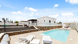 A pool area with white lounge chairs and a stone wall. - Villa doña Isidora - VipVipVillas (Teguise)