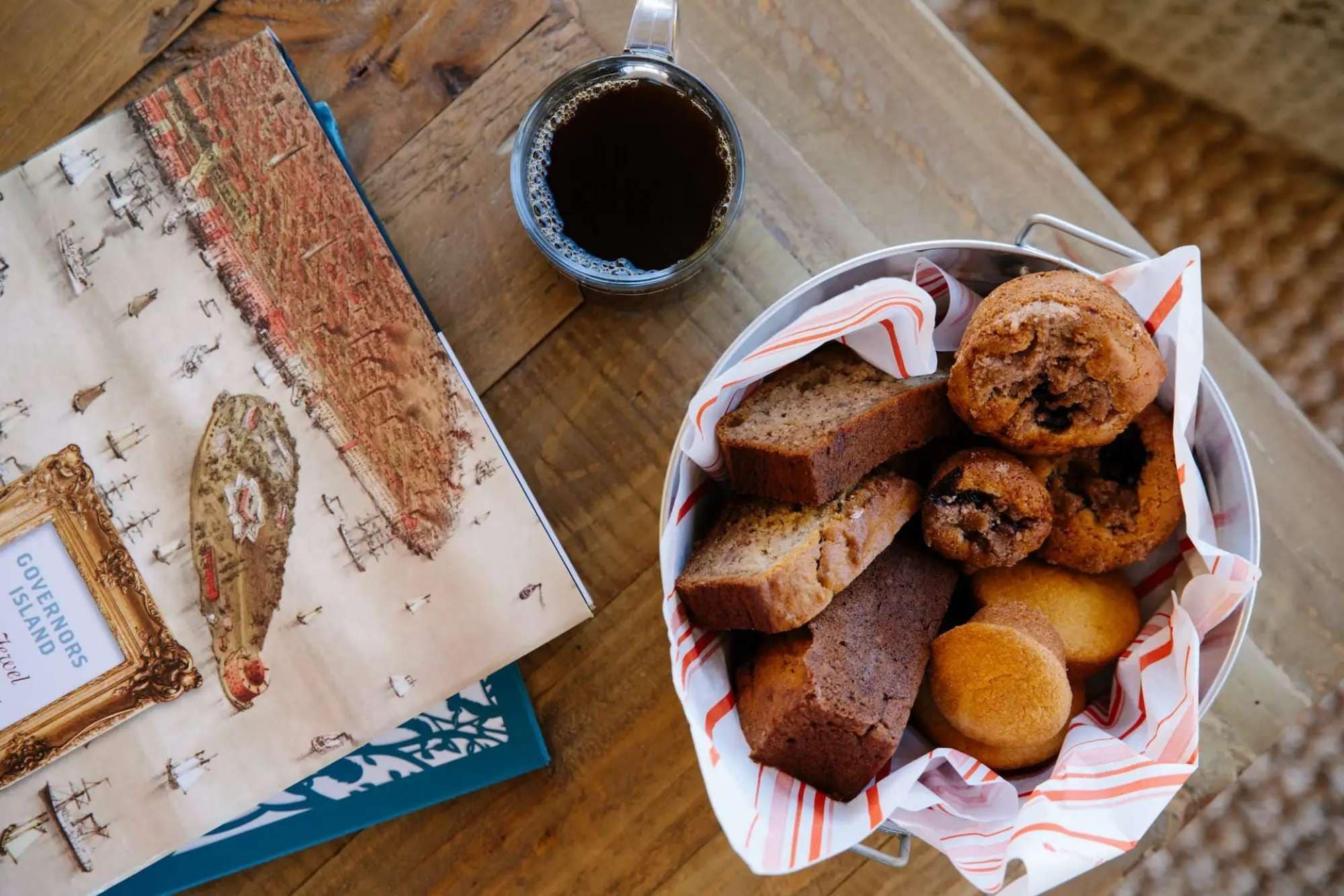 Ett bord med en korg full av muffins och en kopp kaffe, en inramad karta och en bok med titeln ”GOVERNORS ISLAND”.