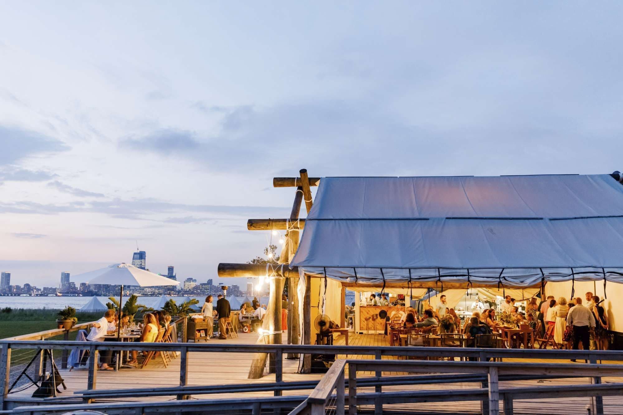 Un restaurant situé au bord de l’eau, avec une terrasse, une vue sur les gratte-ciel de la ville en arrière-plan et une grande structure en forme de tente.