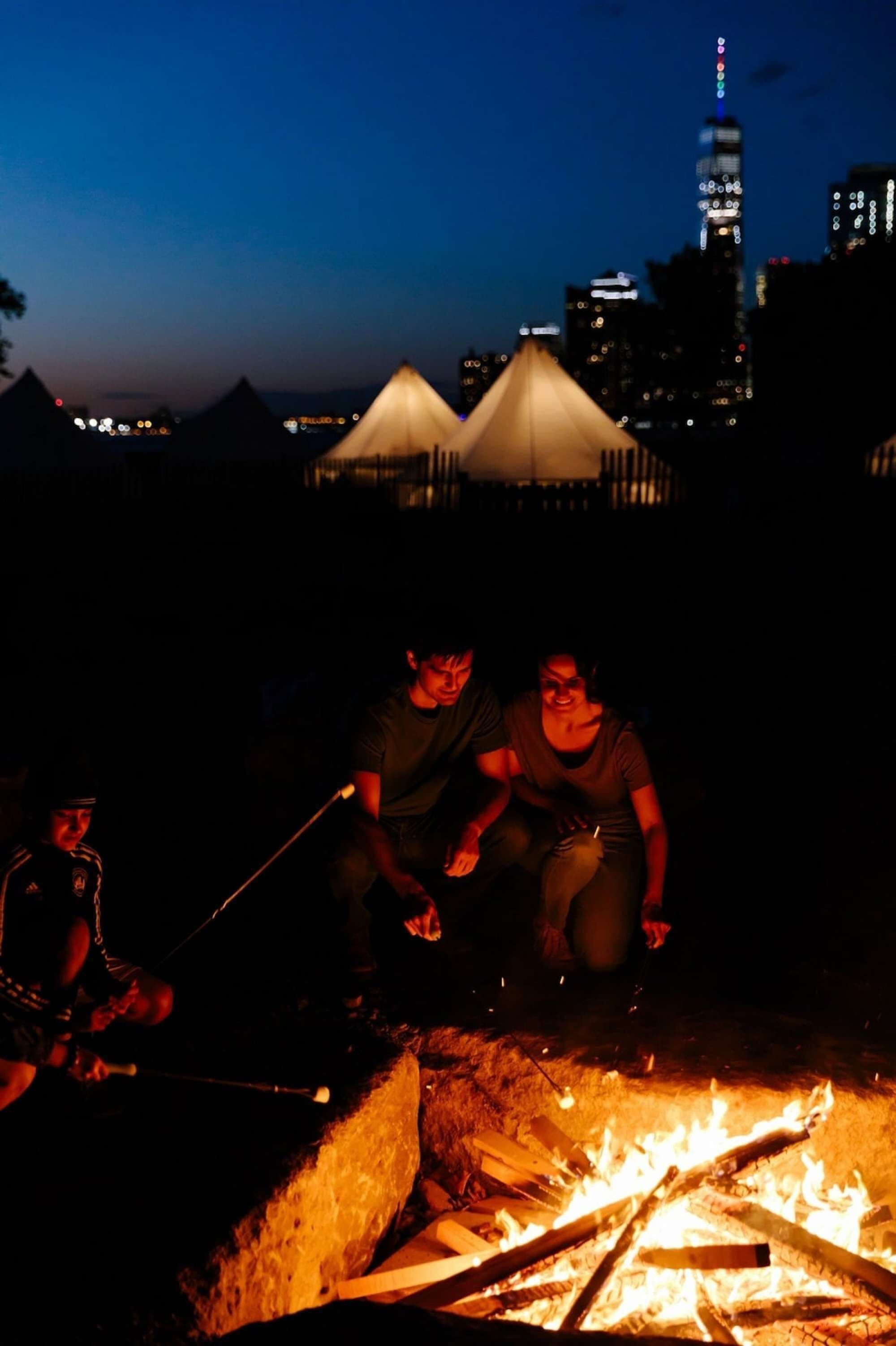 Un groupe de personnes est réuni autour d’un feu de camp la nuit, avec la silhouette d’une ville en arrière-plan.