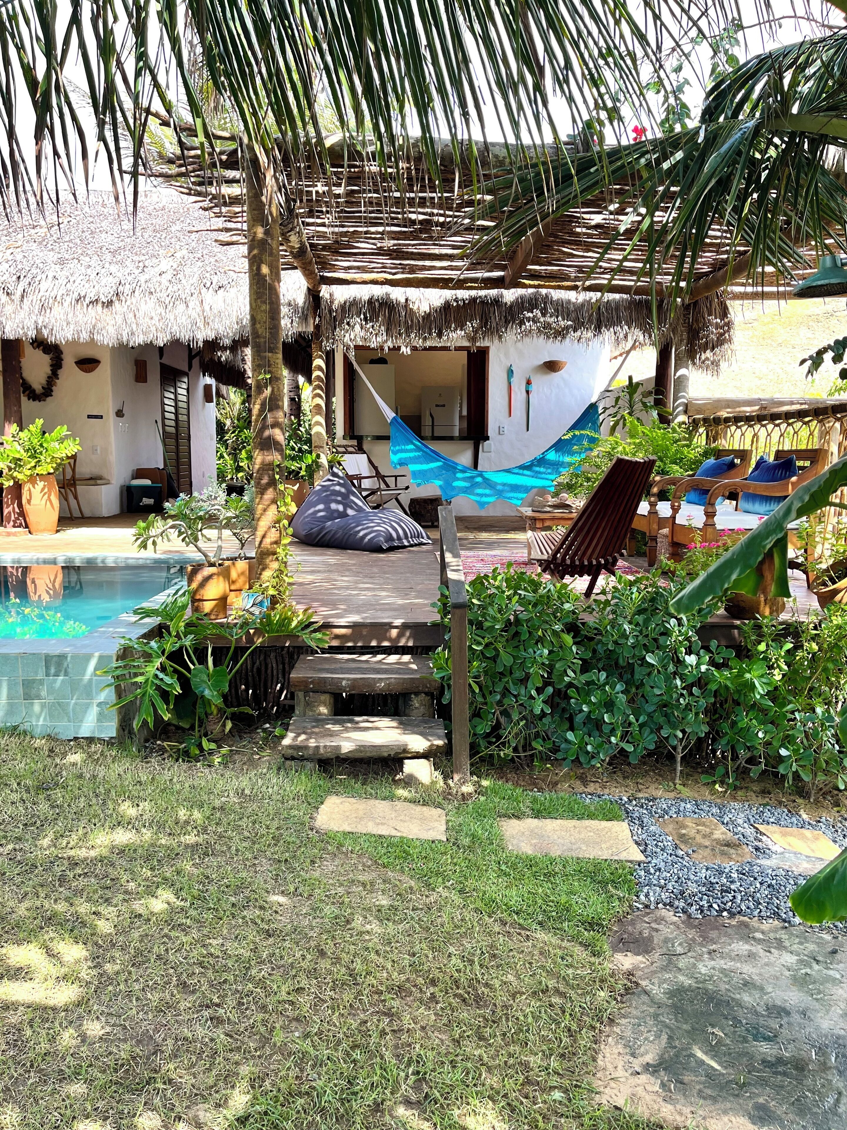 Un espacio al aire libre con techo de paja, una hamaca, una terraza de madera y una piscina.