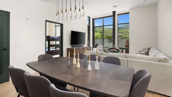 Un espace repas moderne avec une table en bois foncé, des chaises recouvertes d’un tissu gris et un téléviseur en arrière-plan.