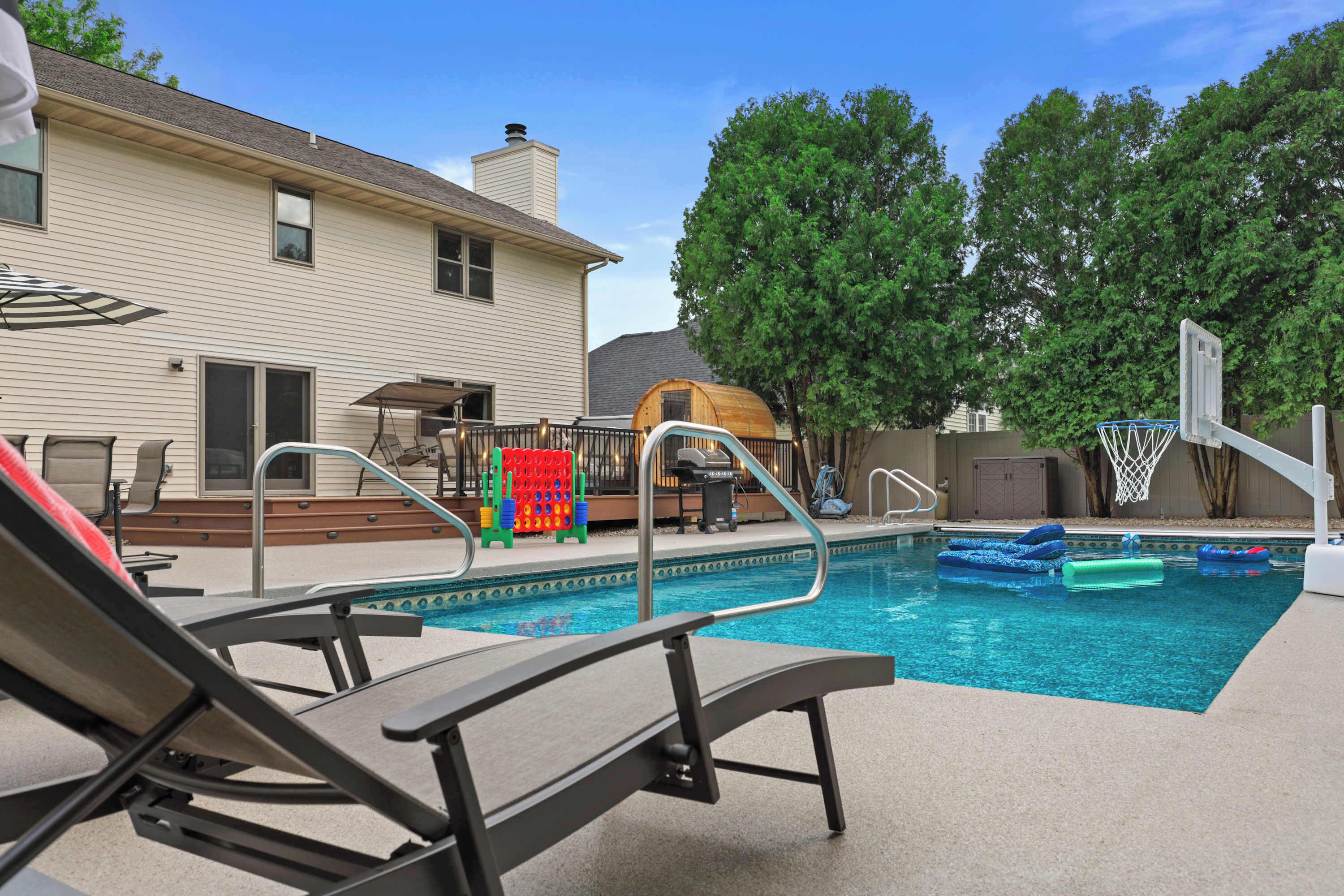 A backyard with a swimming pool, lounge chairs, a basketball hoop, and a wooden playhouse.