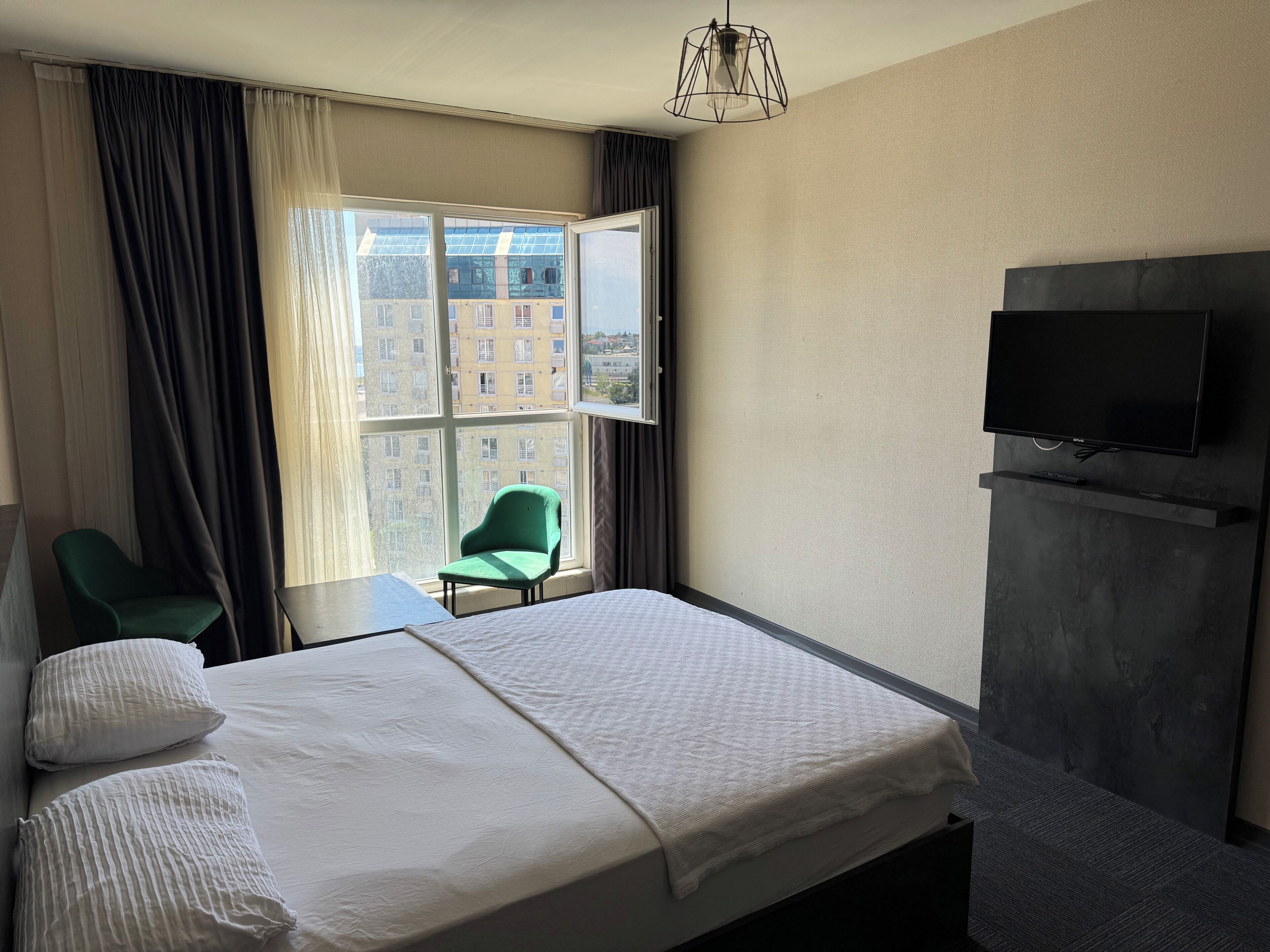 Habitación de hotel con cama, una silla verde, una mesita pequeña, televisor montado en la pared y ventana con cortinas.