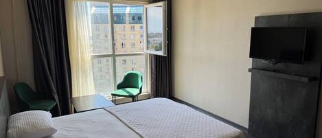 Habitación de hotel con cama, una silla verde, una mesita pequeña, televisor montado en la pared y ventana con cortinas.