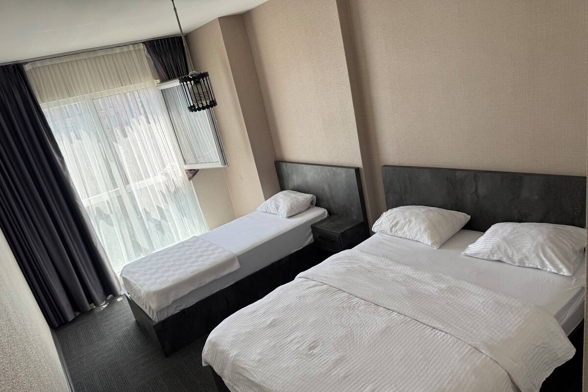 Habitación de hotel con dos camas, un ventanal con cortinas, una mesita de noche y una lámpara colgante.