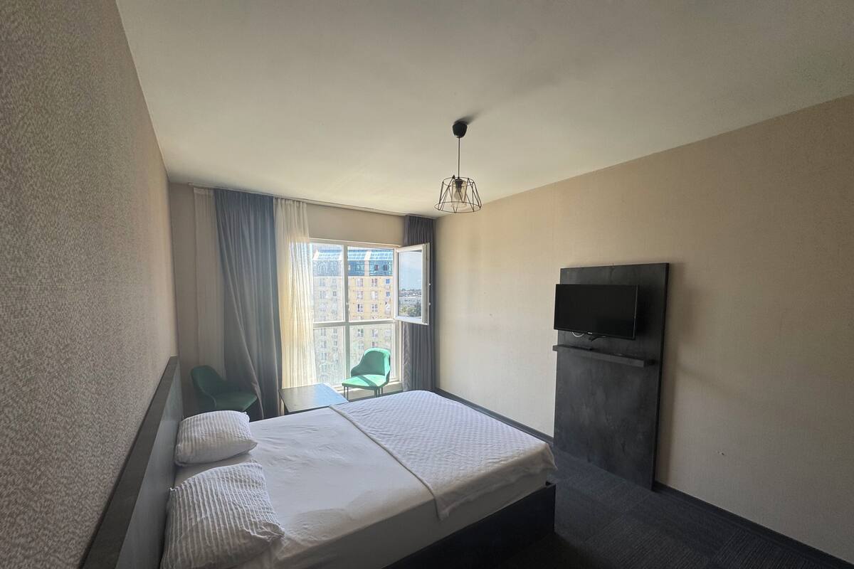 Una habitación de hotel con cama, televisor montado en la pared, una silla y una ventana con cortinas.