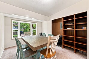 A dining room with a wooden table and chairs, a window with a view of greenery, and a built-in bookshelf.