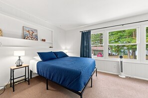 Ein Schlafzimmer mit einem Bett, einem Nachttisch, einer Lampe, einem Fenster mit Vorhängen und Blick ins Freie.