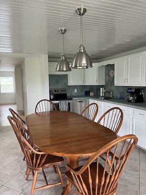 A dining area with a wooden table and chairs, a kitchen in the background, and pendant lighting.