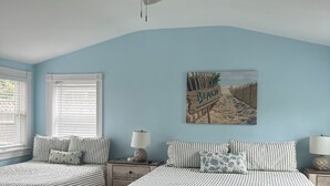 Une chambre avec deux lits, un ventilateur de plafond, une décoration murale sur le thème de la plage et une fenêtre avec des stores.