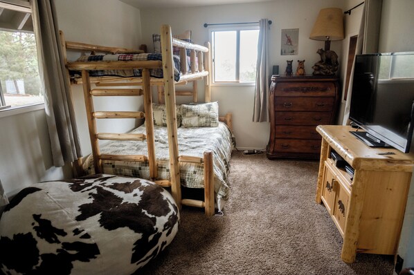 Una cama litera con estructura de madera y una cama individual con manta escocesa. Hay un aparador de madera con un televisor encima, una ventana con cortinas y un sillón de felpa con estampado de vaca.